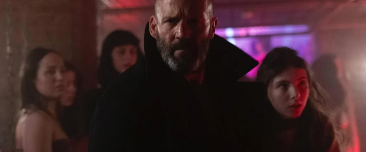 Jason Statham em cena do filme Missão Refúgio (2026) ao lado da jovem Jessie em ambiente iluminado por neon
