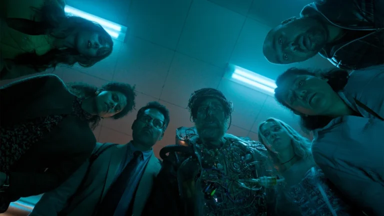Grupo de personagens observa homem com implantes tecnológicos em cena de Boa Sorte, Divirta-se, Não Morra