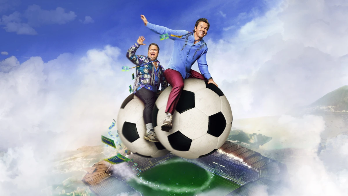 Cena promocional de Bola pra Cima com Mark Wahlberg e Paul Walter Hauser sentados em bolas de futebol sobre um estádio