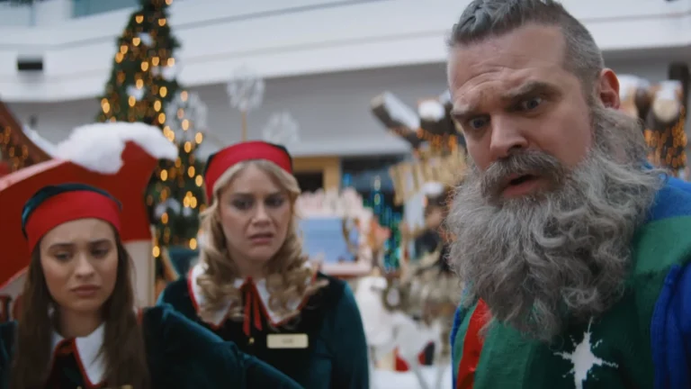 Papai Noel Volta Armado (e Casado) em Noite Infeliz 2 1 Papai Noel, interpretado por David Harbour, observa algo com expressão de choque enquanto duas ajudantes natalinas aparecem ao fundo em cena de "Noite Infeliz 2".