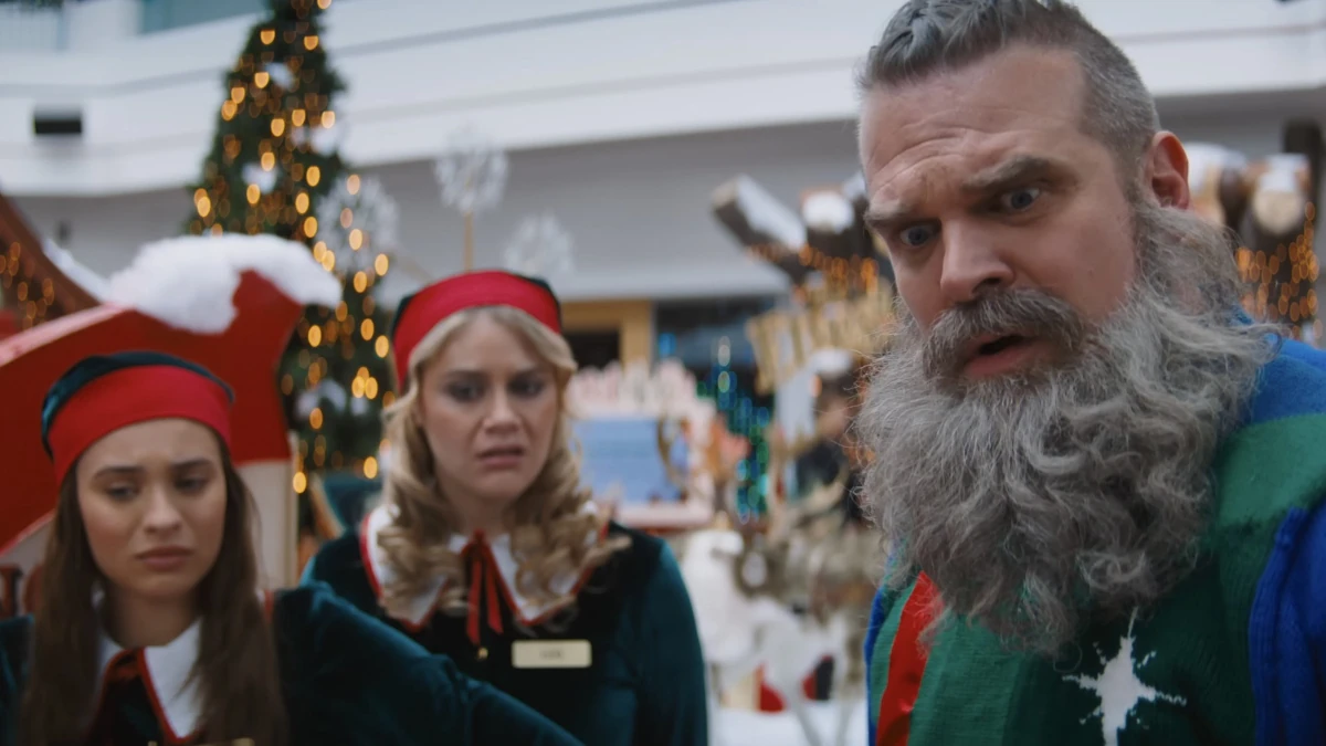 Papai Noel Volta Armado (e Casado) em Noite Infeliz 2 1 Papai Noel, interpretado por David Harbour, observa algo com expressão de choque enquanto duas ajudantes natalinas aparecem ao fundo em cena de "Noite Infeliz 2".